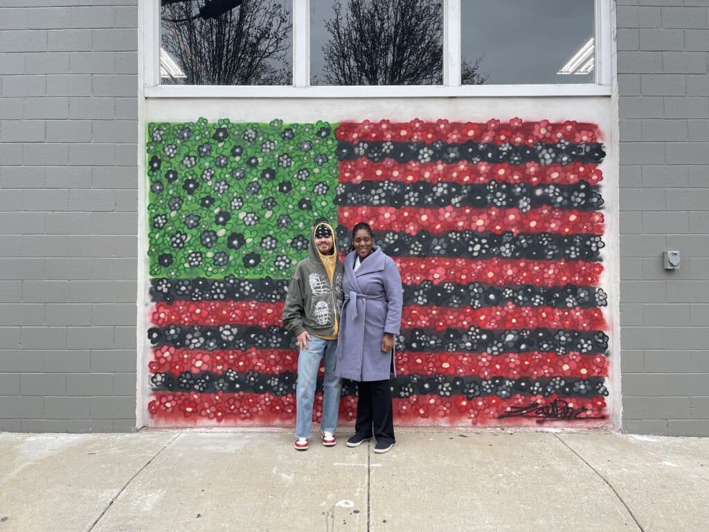 Mural Arist, Jordan Carter (left), and The Heritage Club Founder, Nike John (right), stand in front of Jordan's mural - David Hammons’ African American Flag.